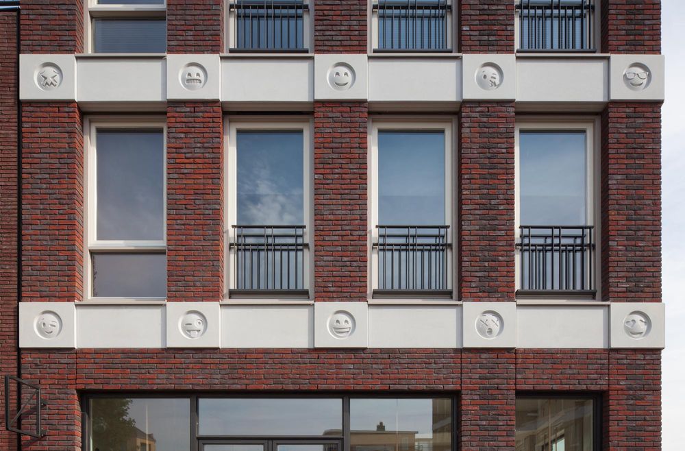 Close up of facade of brick building with face emojis cast in white concrete that run horizontally at the top of each story. 