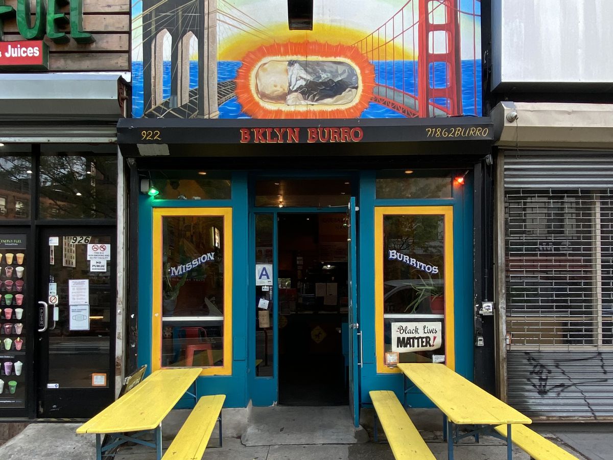 A colorful restaurant with two yellow picnic tables out front and a painted mural of the San Francisco Bridge.