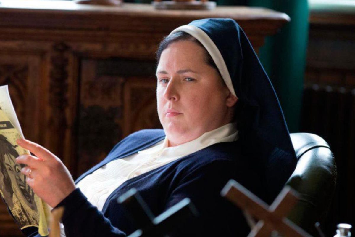 Sister Michael reads a leaflet called “The Habit” while wearing her habit, seated in a chair in Derry Girls.