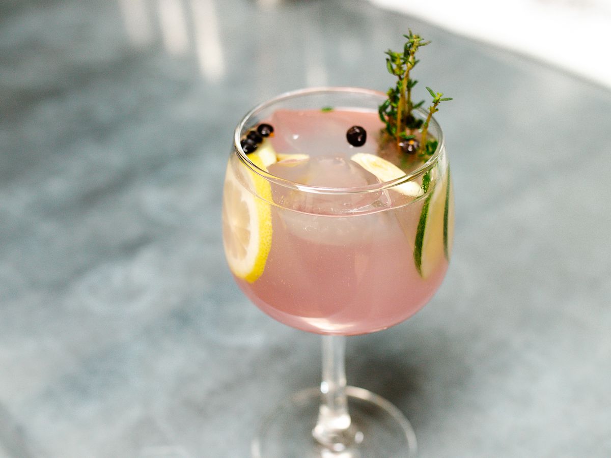 A pink cocktail garnished with lemon, cucumber and herbs, place on a metal bar. 