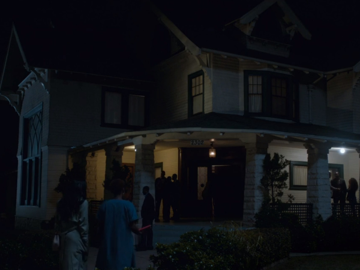 The exterior of a house at night. There are people outside of the house. The house has a wraparound porch and multiple floors.