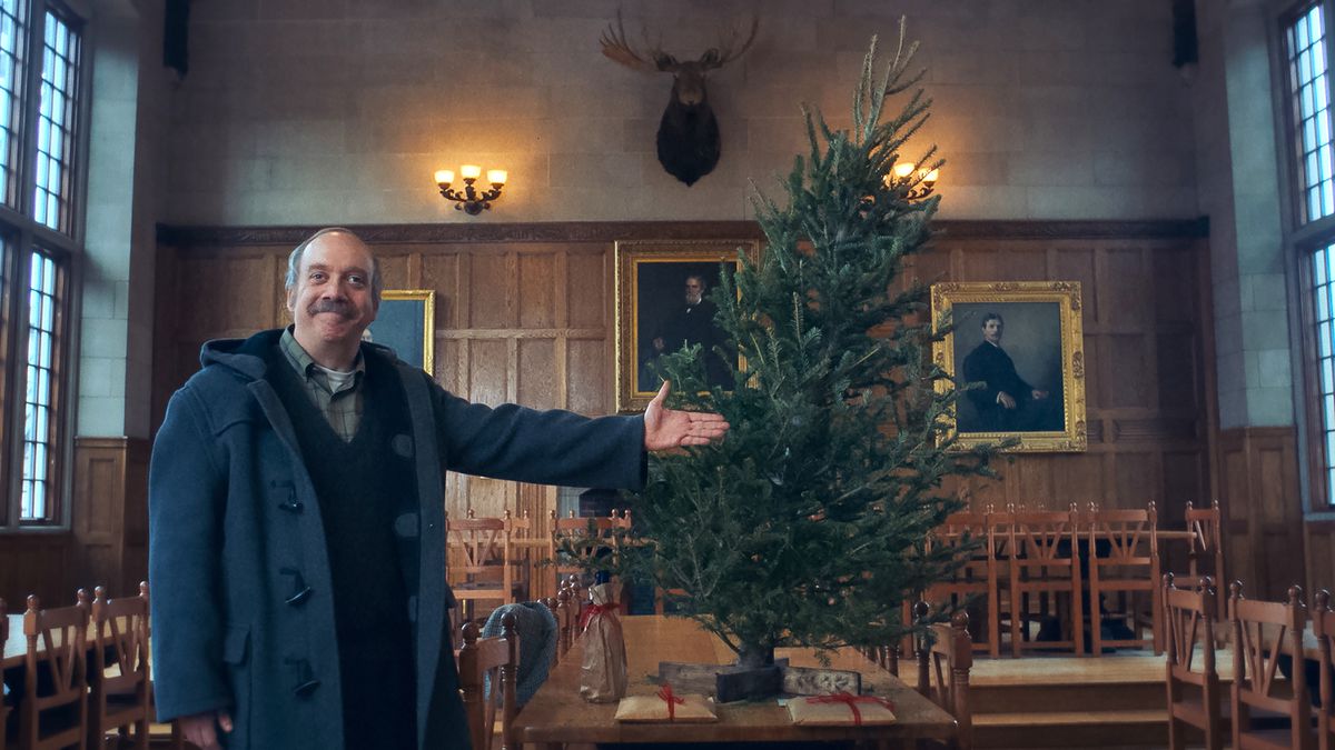 Boarding-school professor Paul Hunham (Paul Giamatti) smiles proudly while indicating the naked, listing pine tree he bought for Christmas, positioned on a table in the middle of a cluttered boarding-school room in The Holdovers