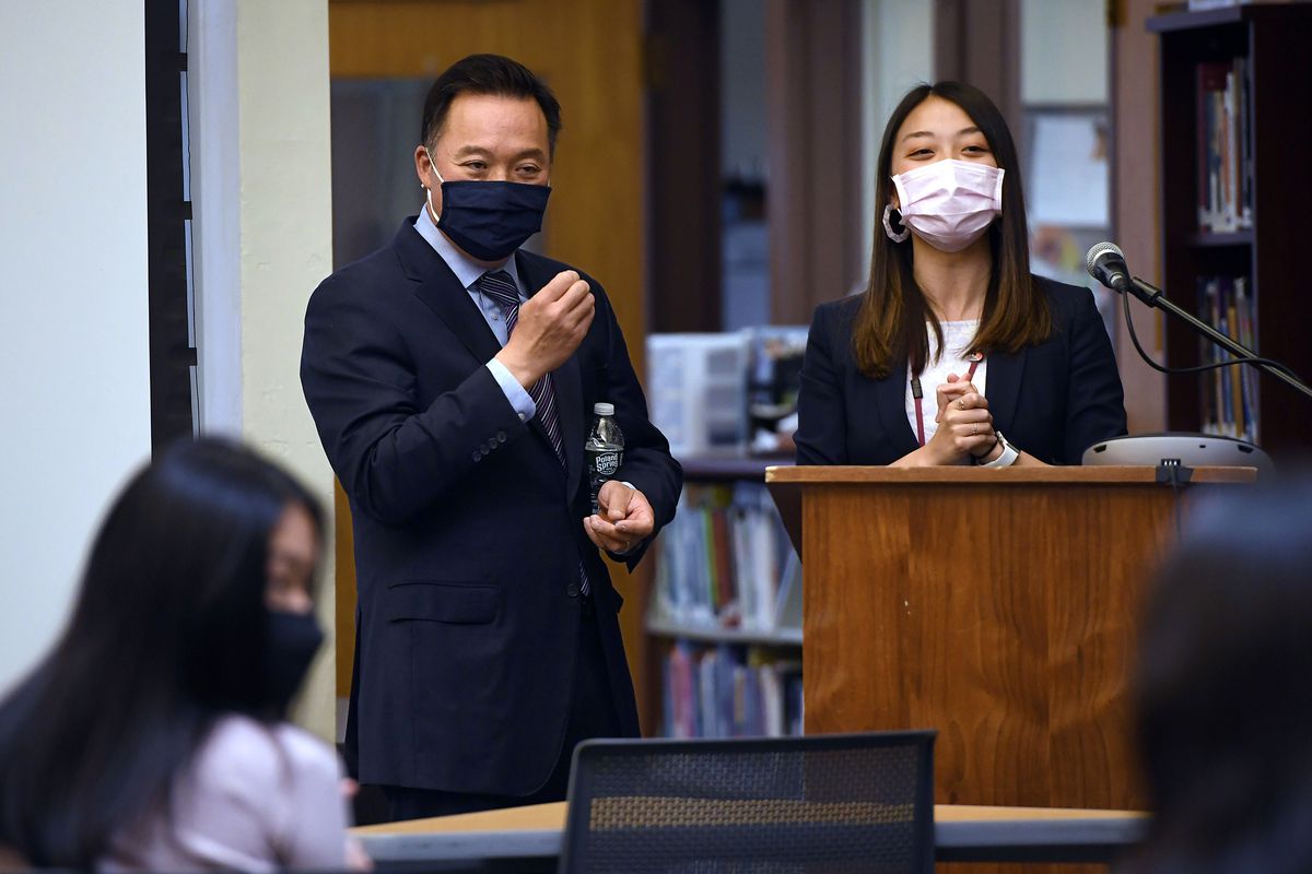 Tong and Tan, both in navy suits, and each in a surgical mask, stand behind a podium. Tan has her hands folded; Tong is gesturing with his hand, appearing to be answering a question.