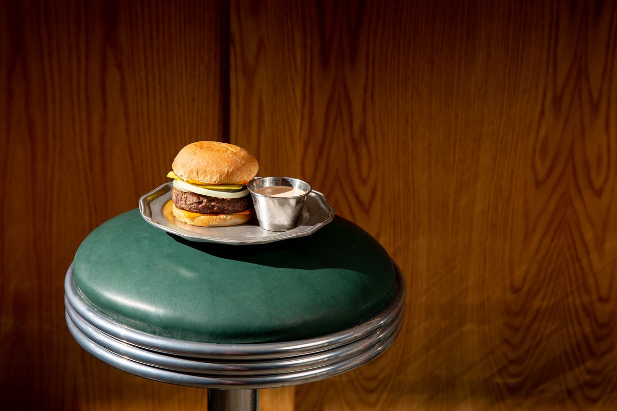 A traditional burger with gravy on the side on a silver platter sits on a green leather diner stool.