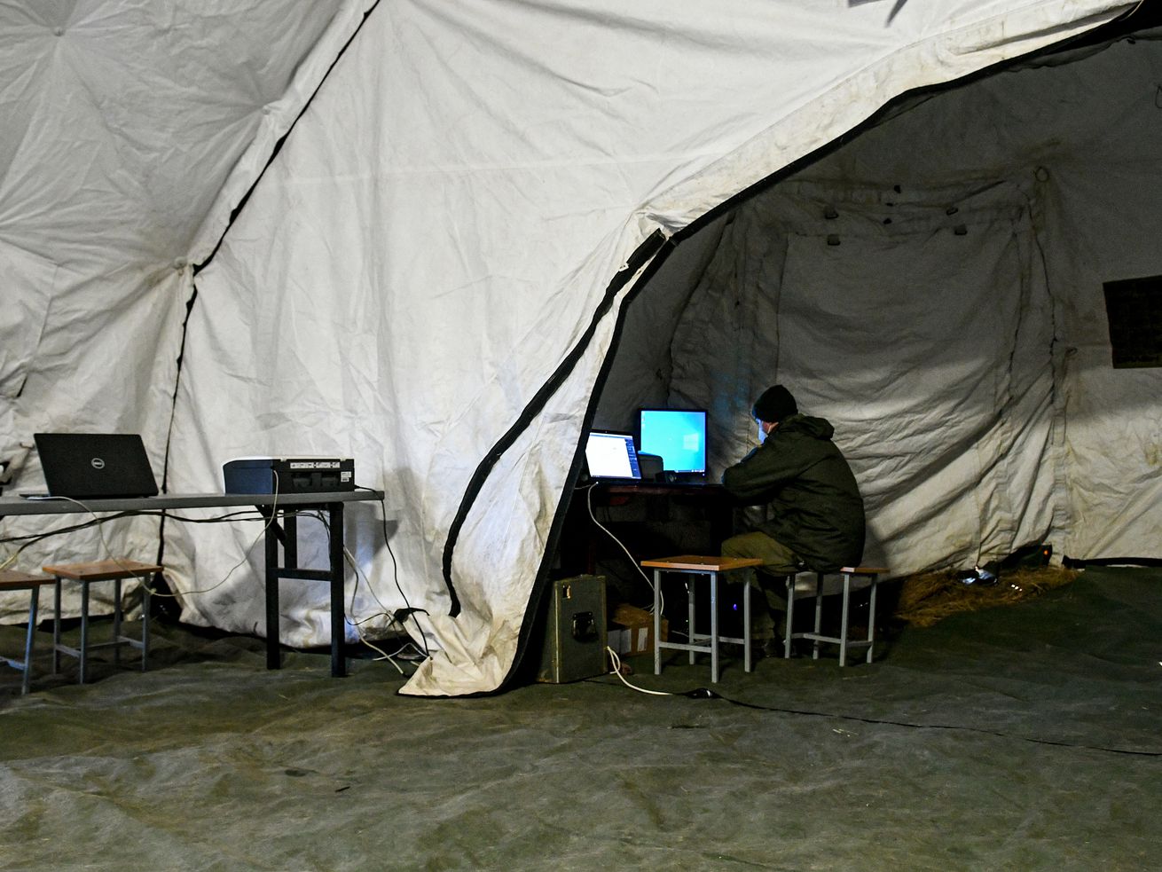 A person inside a large military tent sits at a desk with a computer. 