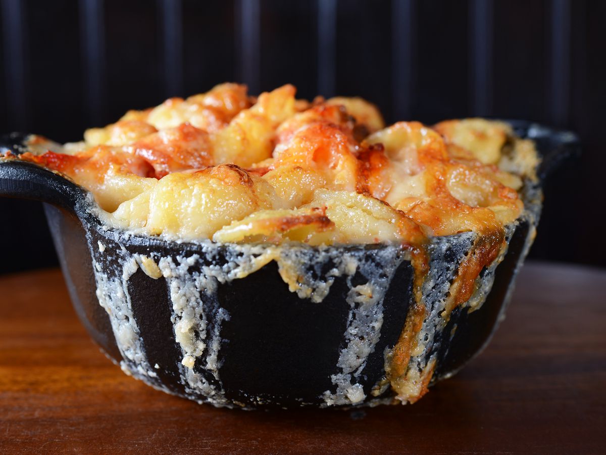 The main focus in this picture is a black bowl with lobster mac and cheese. What gets your attention is the cheese has overflowed the bowl and melted down the sides. Out of focus, in the background is the seat back of a booth in the restaurant.