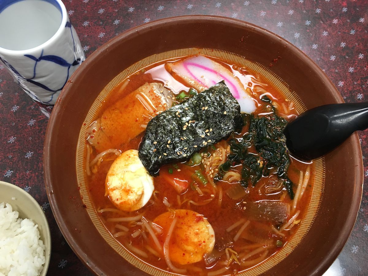 A brown bowl of ramen with a bright red broth, boiled eggs, and nori. 
