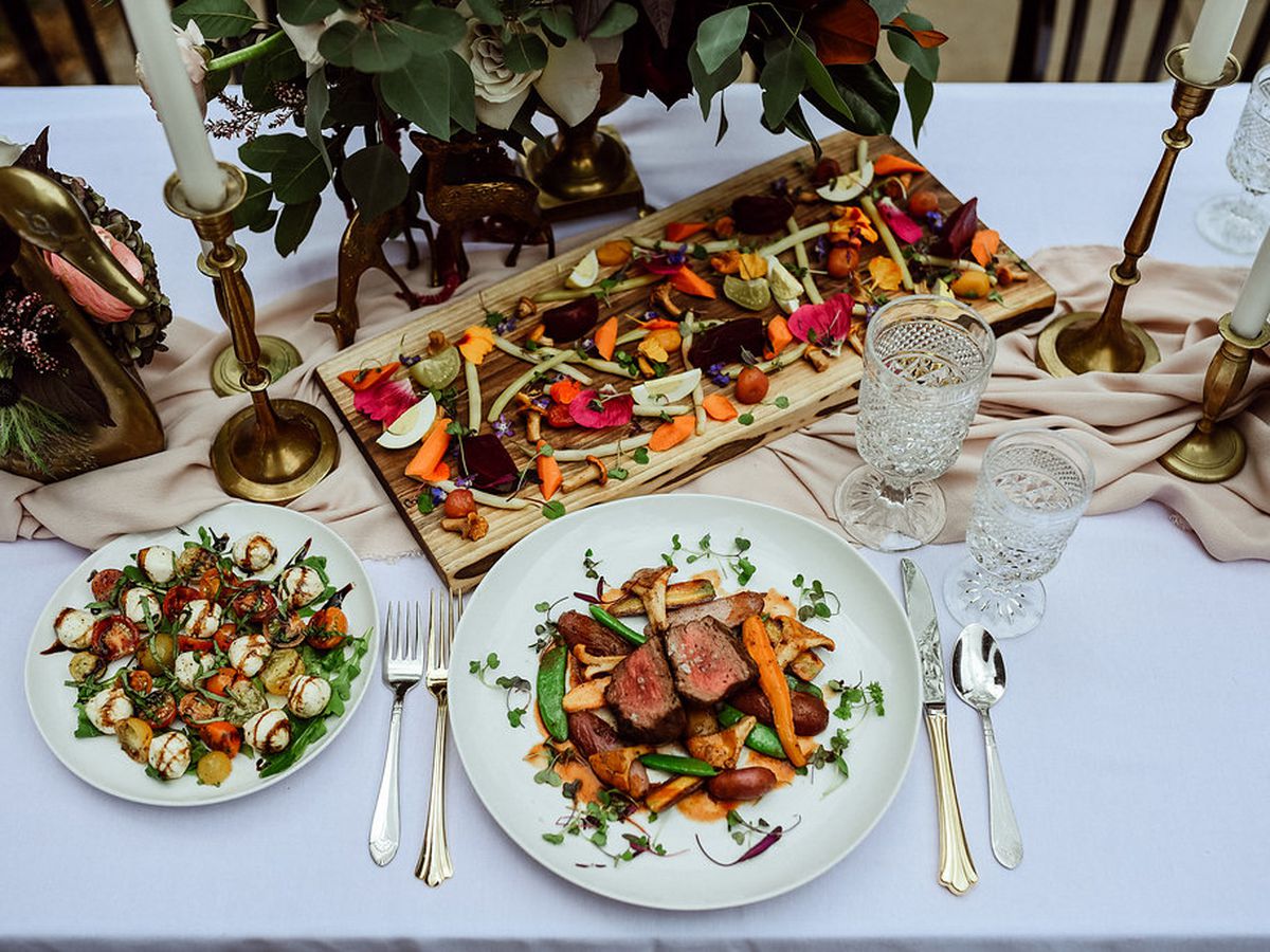 A spread of meat and vegetable plates on a white tablecloth with brass candlesticks and greenery. 