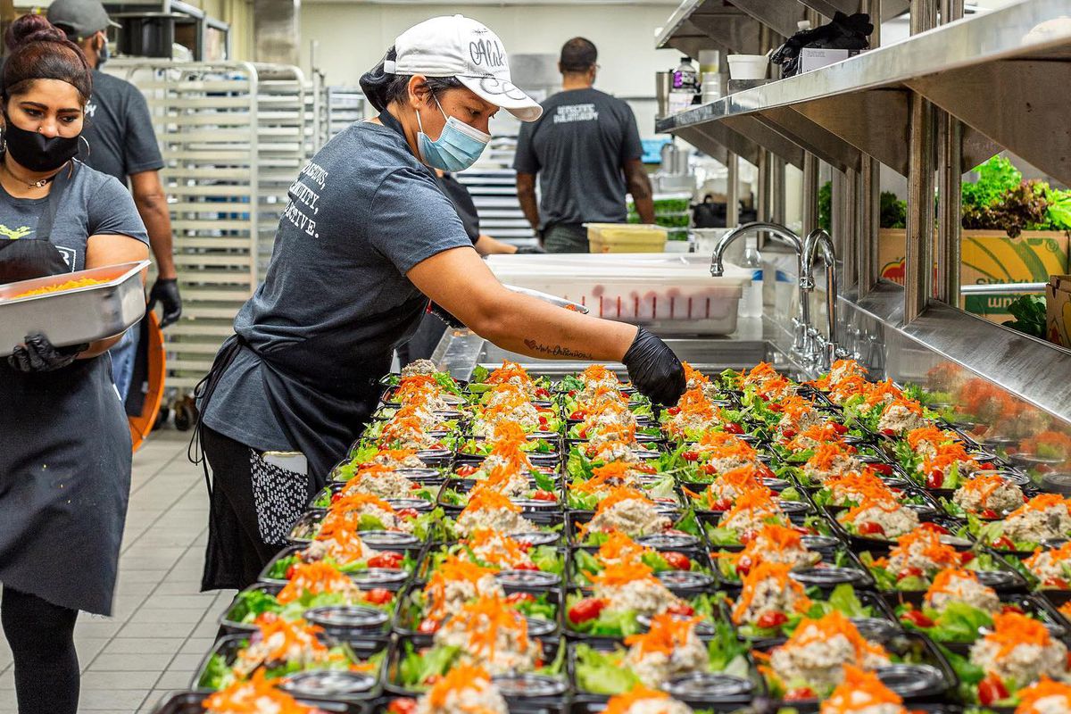 a group of masked kitchen workers garnishes a massive line of meals intended to be handed out for christmas