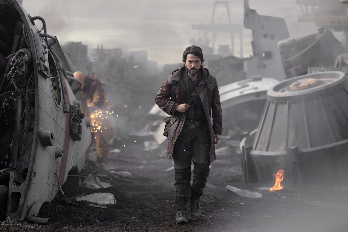 Diego Luna as Cassian Andor walks through a field of scrap, with machines broken and on fire.
