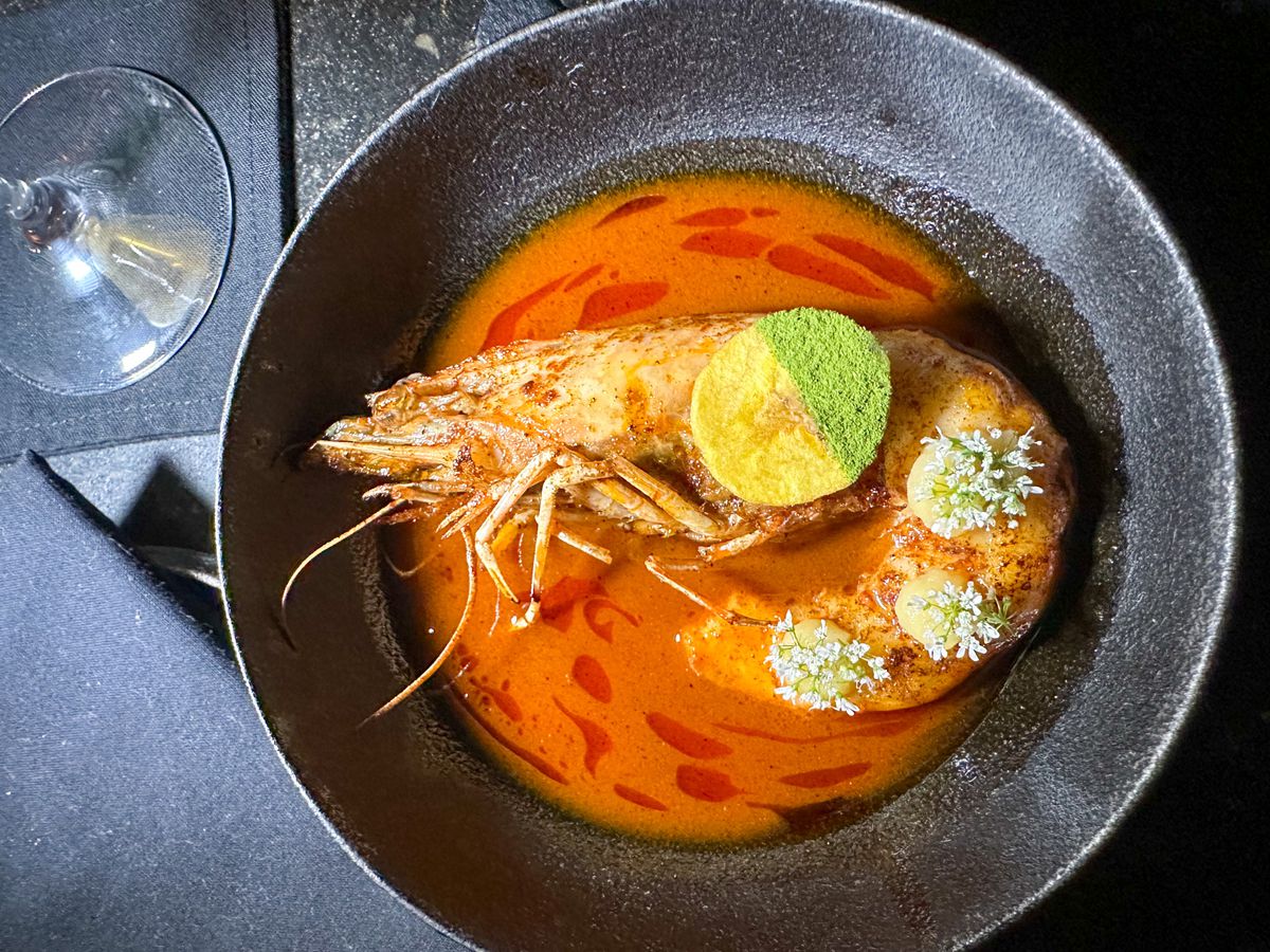 A whole prawn in red broth, with clumps of emulsion for garnish and a lemon wedge dusted with green powder.
