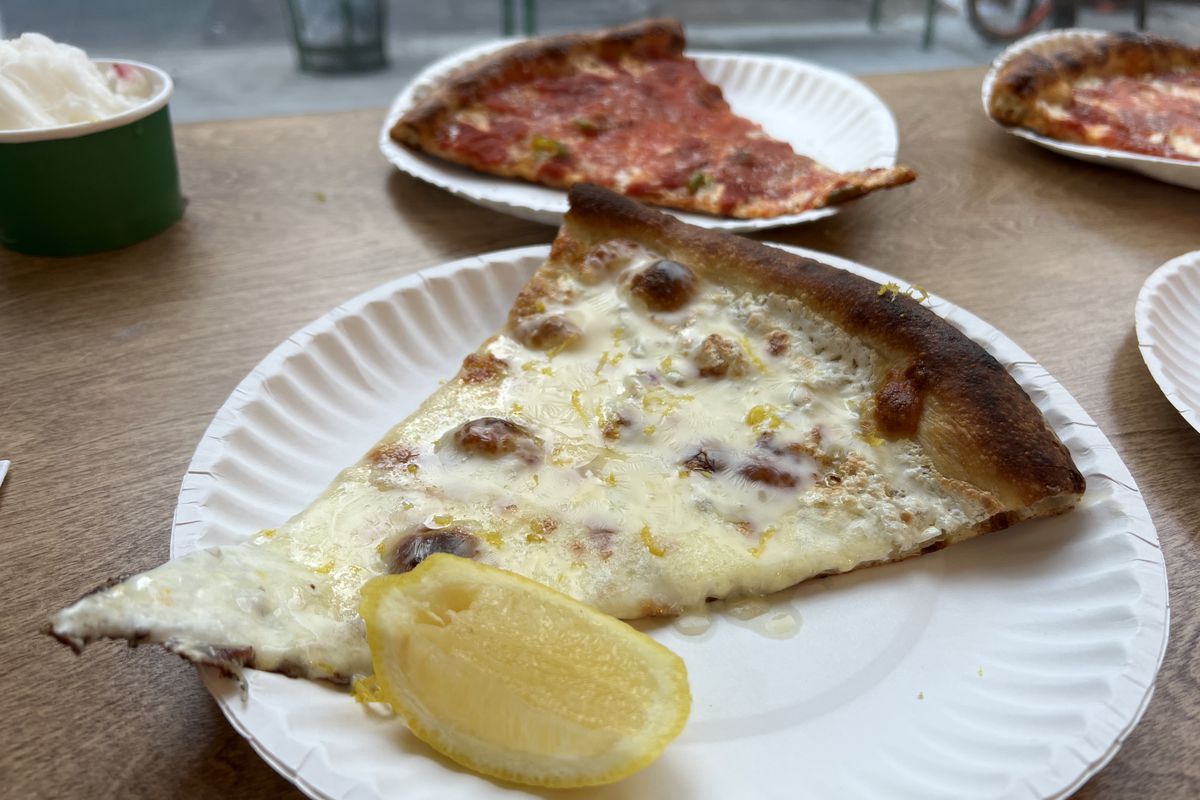 A white slice of pizza, sprinkled with lemon zest, sits on a paper plate, with a lemon wedge on the site; cheese slices are visible in the background