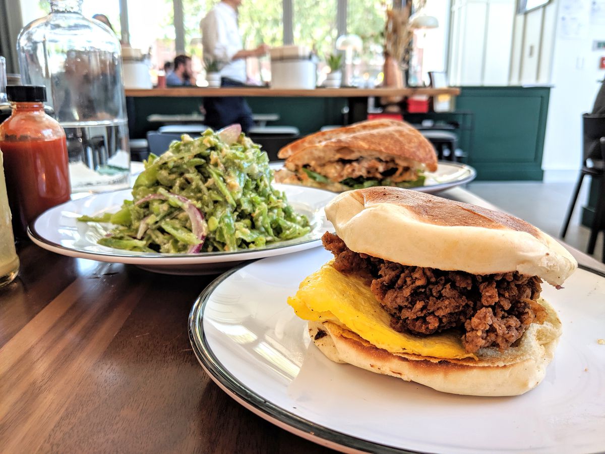 A fried chicken sandwich served on a Portuguese bun is in the foreground of the photo, with a stacked-high sugar snap pea salad and a roasted cauliflower sandwich in the background. Shot at Shy Bird in Cambridge.