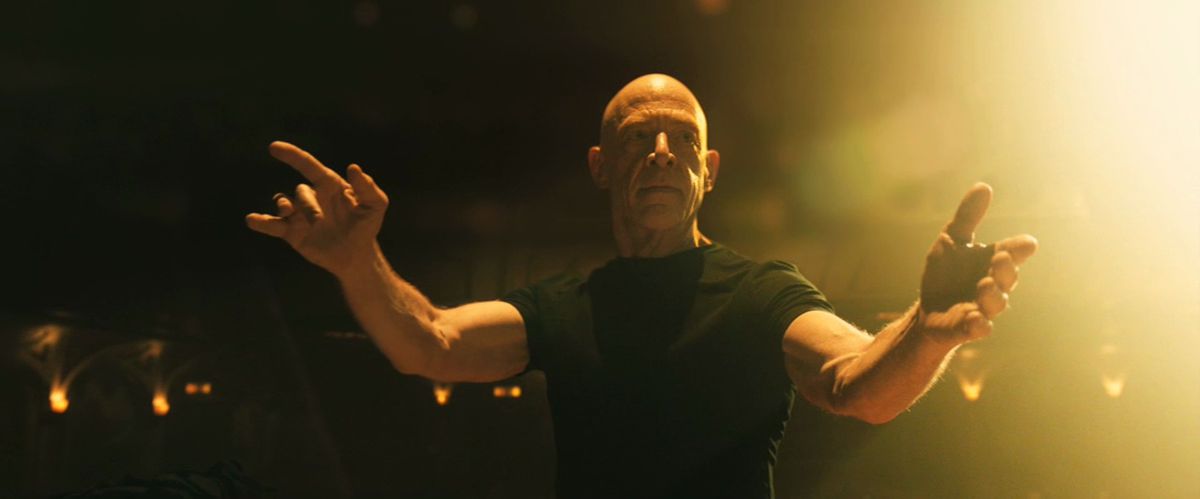 A bald man in a black short-sleeve t-shirt standing on a dark stage with his hands outstretched.