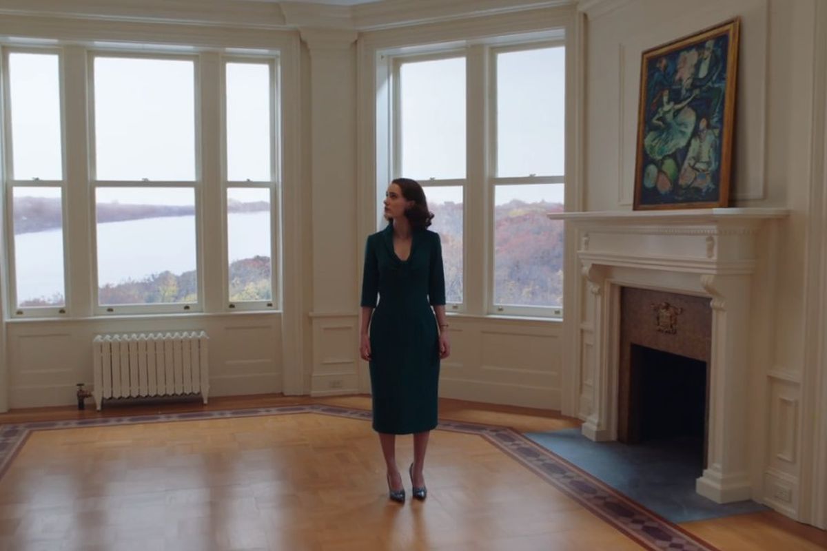 A woman stands in a bright living room with windows overlooking a river.