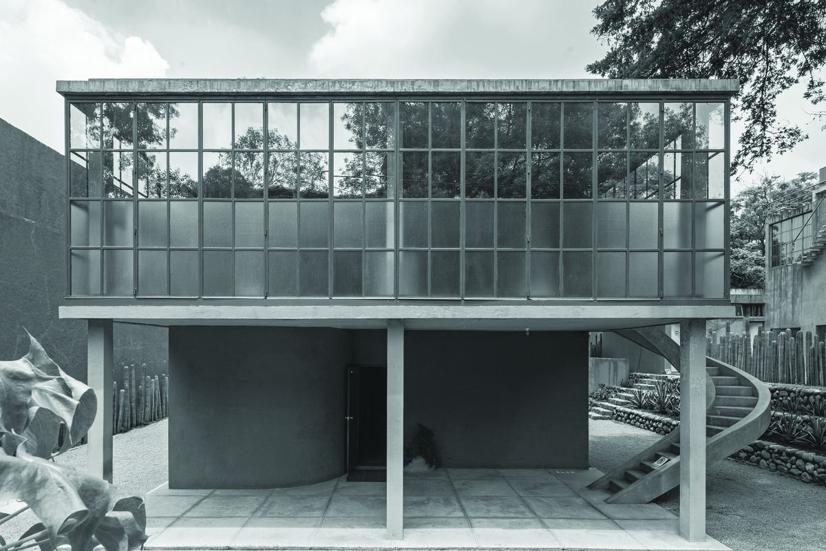 Black and white photo of boxy glass-walled structure set atop a concrete base on stilts.