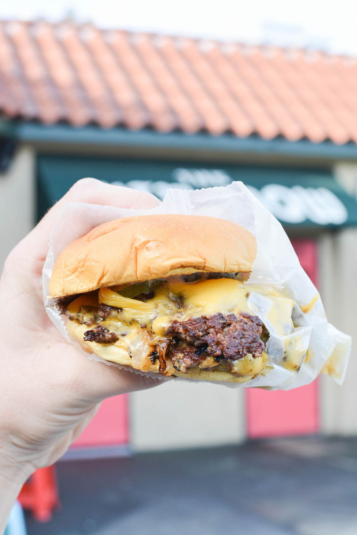 A hand holds a smashed burger with pickles in front of a green restaurant awning.