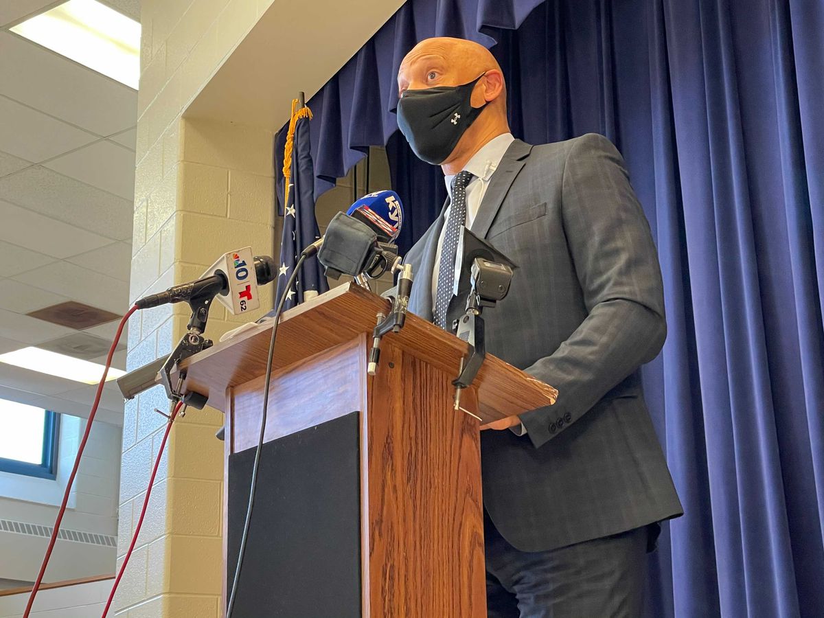 William Hite wearing a face mask standing at a podium with microphones.