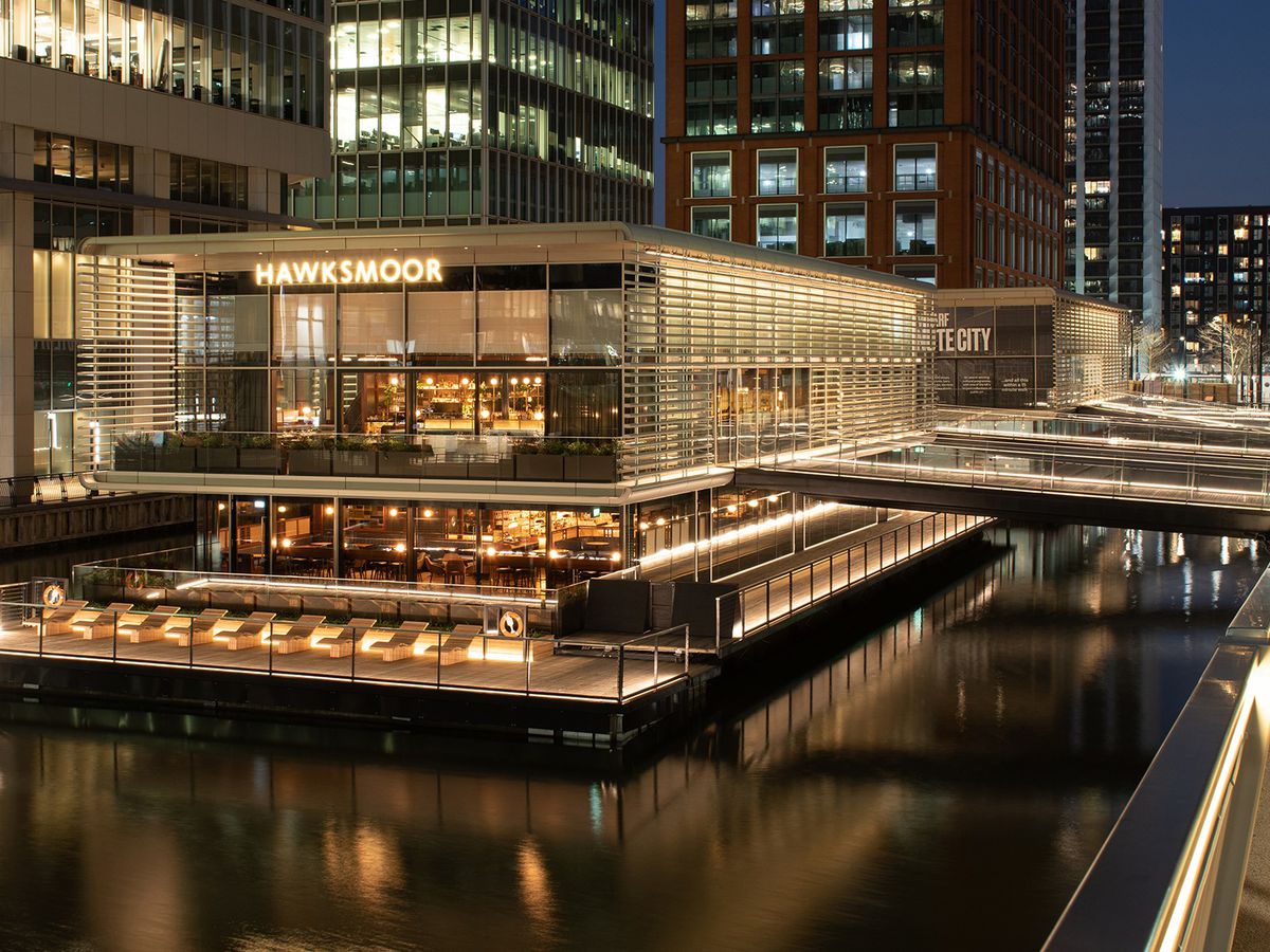 A floating restaurant and bar on three levels in Canary Wharf, with skyscrapers all around, at night.