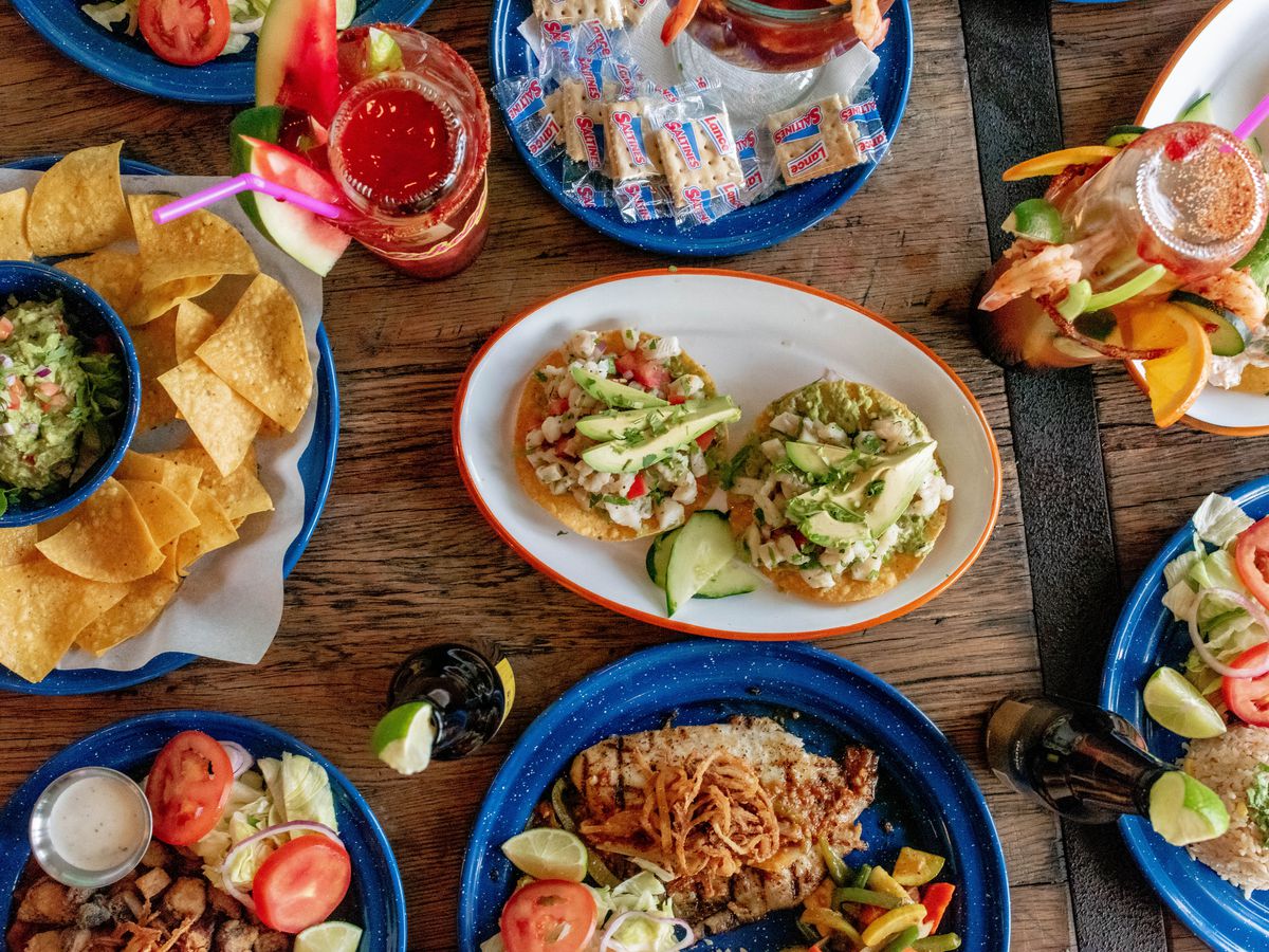 Various Northern Mexican dishes at La Pesca Market, presented on a wooden tabletop, all on rich blue plates. From left to right, clockwise: guacamole and tortilla chips, shrimp tostadas, a michelada, a partial plate with rice and freshly sliced tomatoes, a partial plate with chicken.