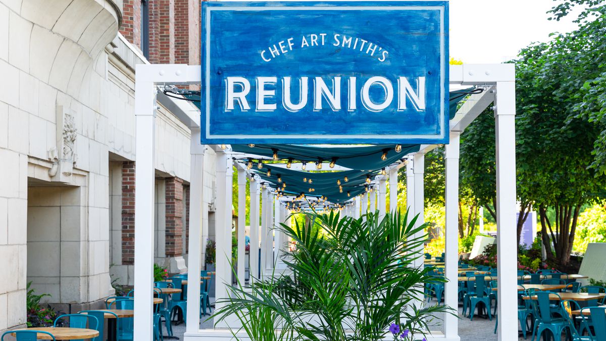 A blue wooden sign in front of Art Smith’s Reunion outside on a sidewalk patio with bushes and patio furniture.
