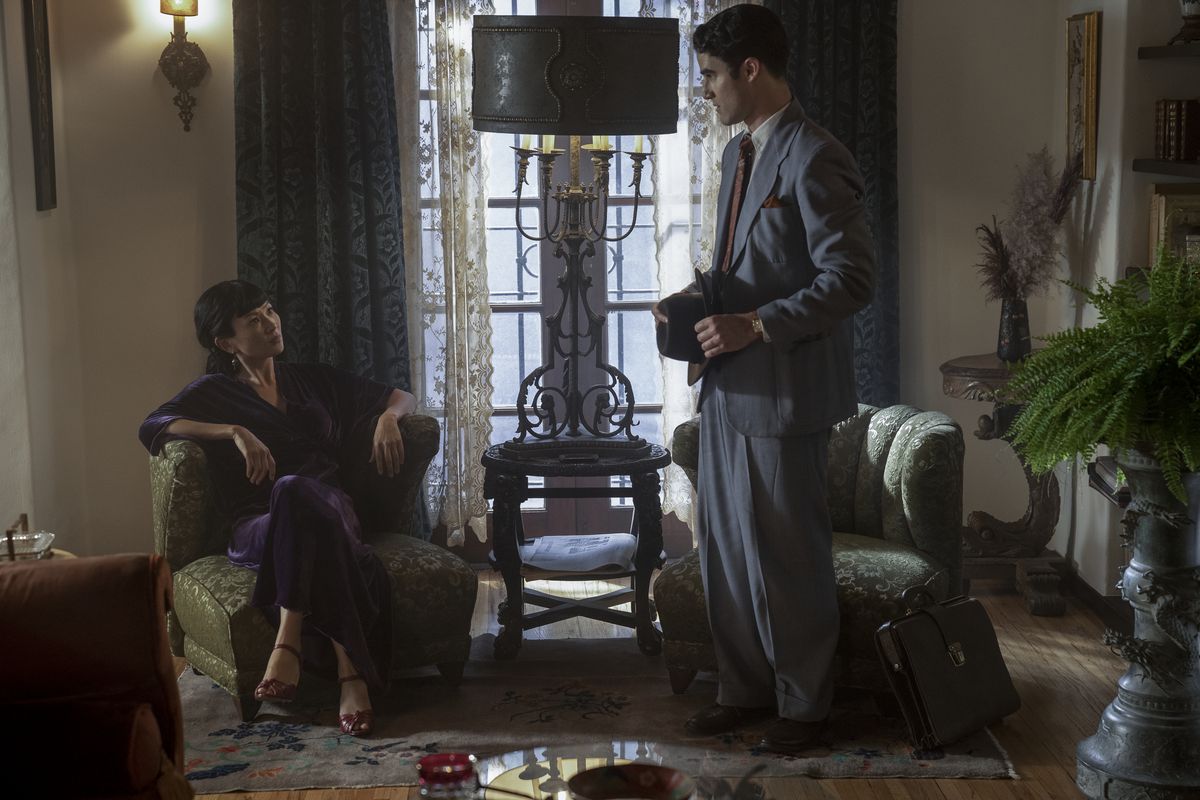 Darren Criss, in a grey tailored suit, stands over a smirking Michelle Krusiec, sitting cross-legged in a plush chair.