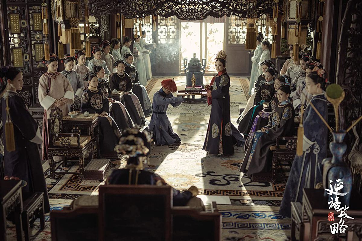A person kneels in front of another, both of whom are surrounded by seated figures in an Chinese palace.