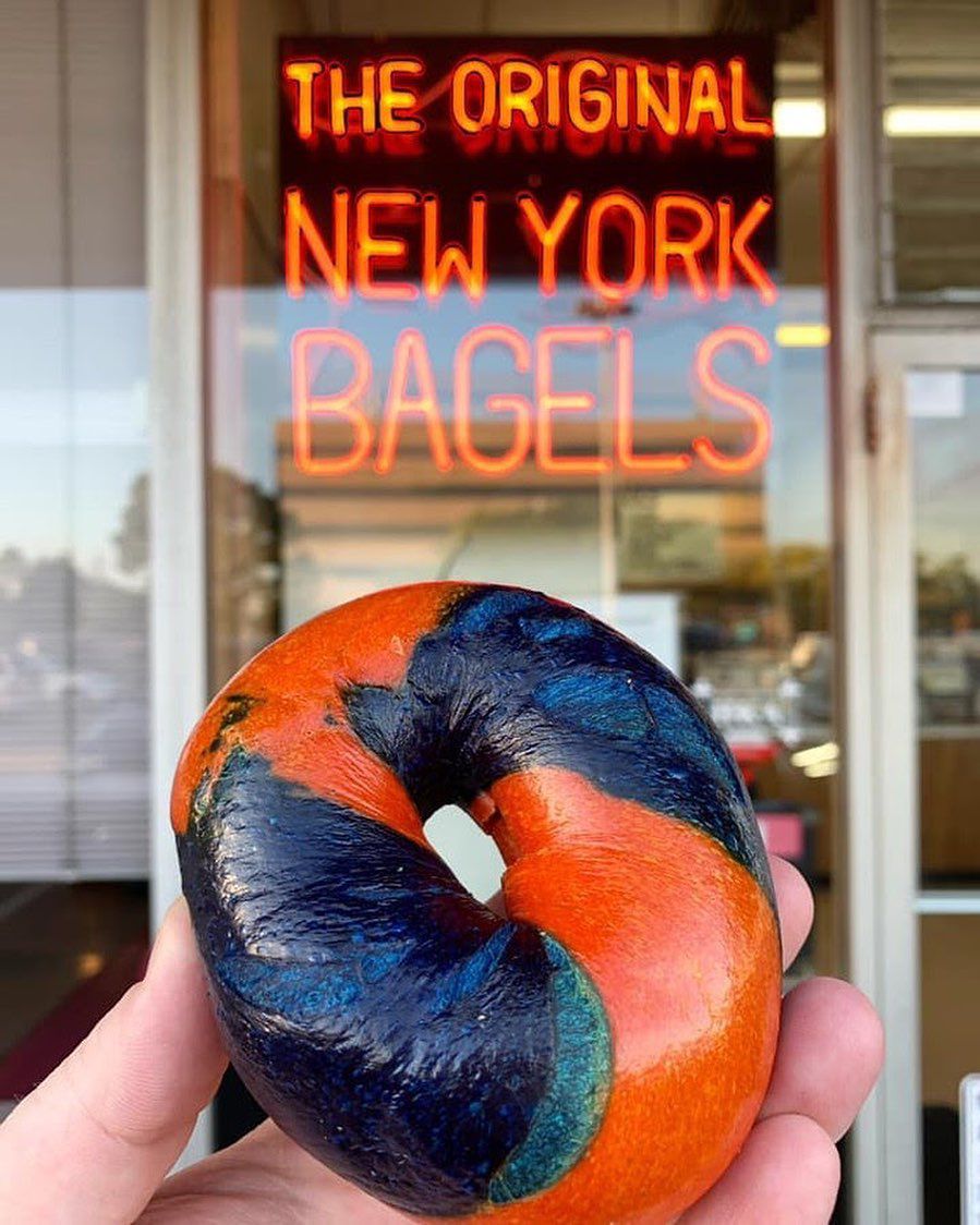 A close-up of a person holding up an orange and blue-swirled bagel in front of a “The Original New York Bagels” sign.