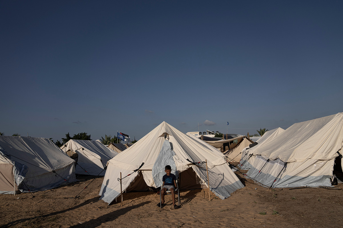 Rows of white tents are erected on the sand. A boy sits alone on a single chair.