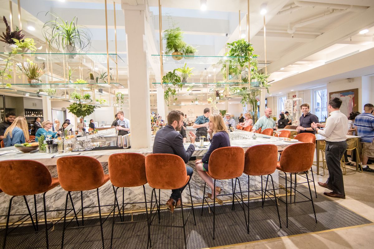 People sit in velvet stools at a marble bar lined with plants on shelves above.