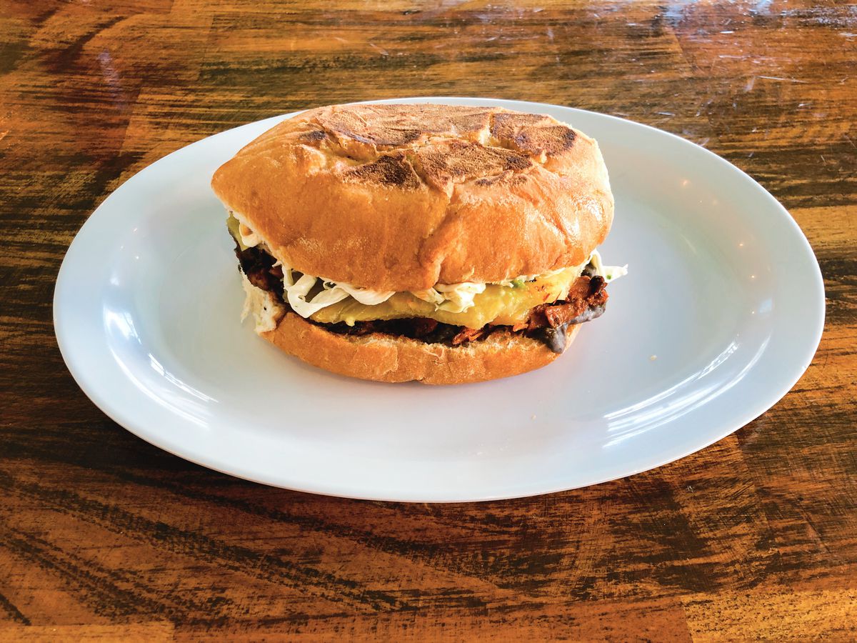 A Mexican torta —&nbsp;a round sandwich a little bigger than a standard hamburger —&nbsp;sits on a white plate on a wooden table.