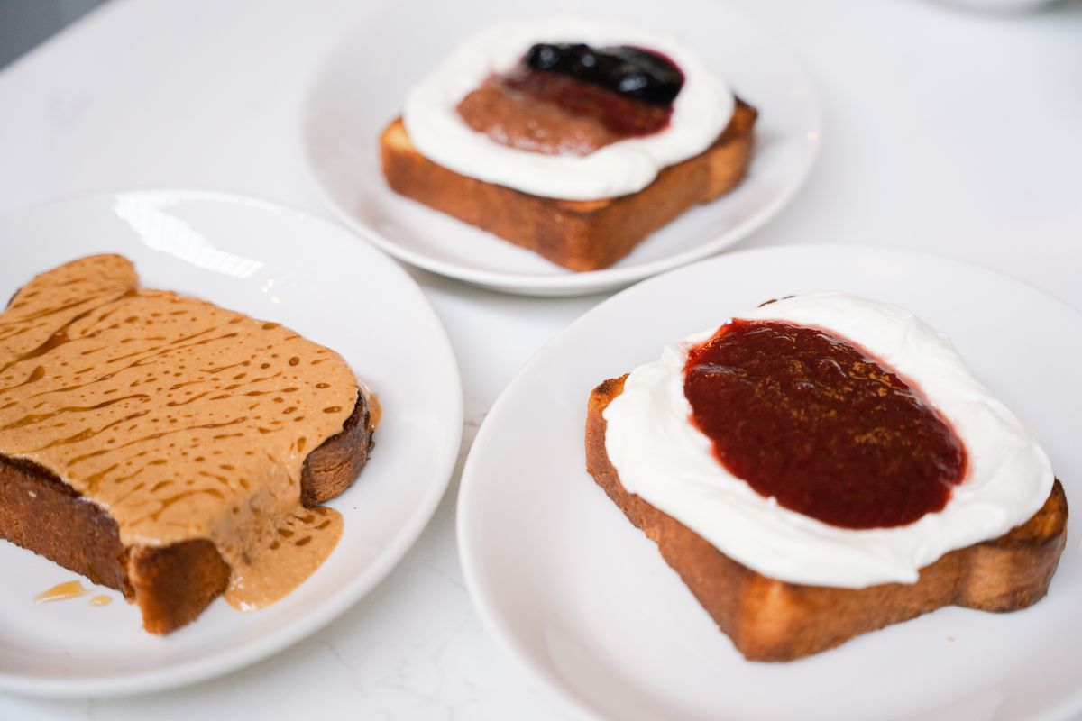 Three toasts sit on white plates; the one to the far left has nut butter, the one to the right has red jam and ricotta; and the one in the back has a mix of different jams with ricotta.