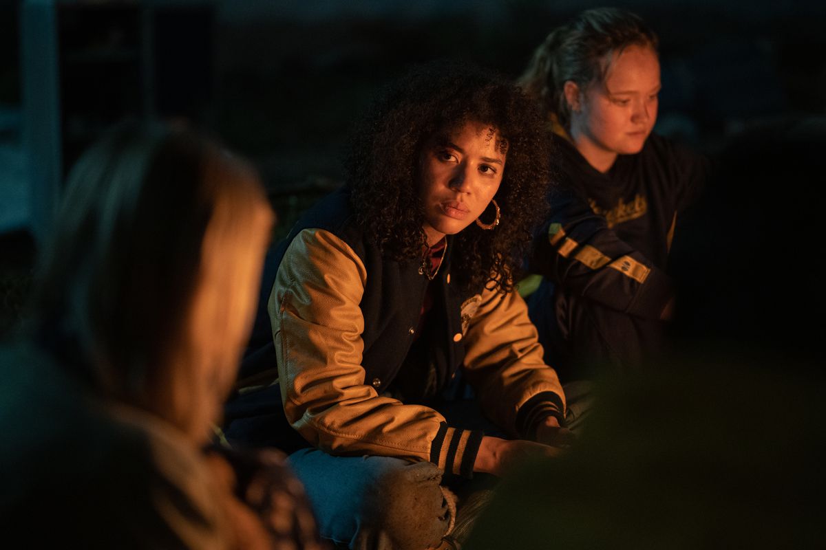 The teenaged version of Taissa, played by Jasmin Savoy Brown, sits next to the fireside in the wilderness, next to her girlfriend Van (played by Liv Hewson)