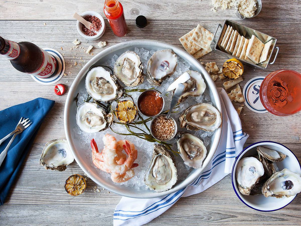 A circular tray of halved oysters and a pile of pink shrimp and ice are surrounded by a little bowl with empty oyster shells, condiments, and crackers.