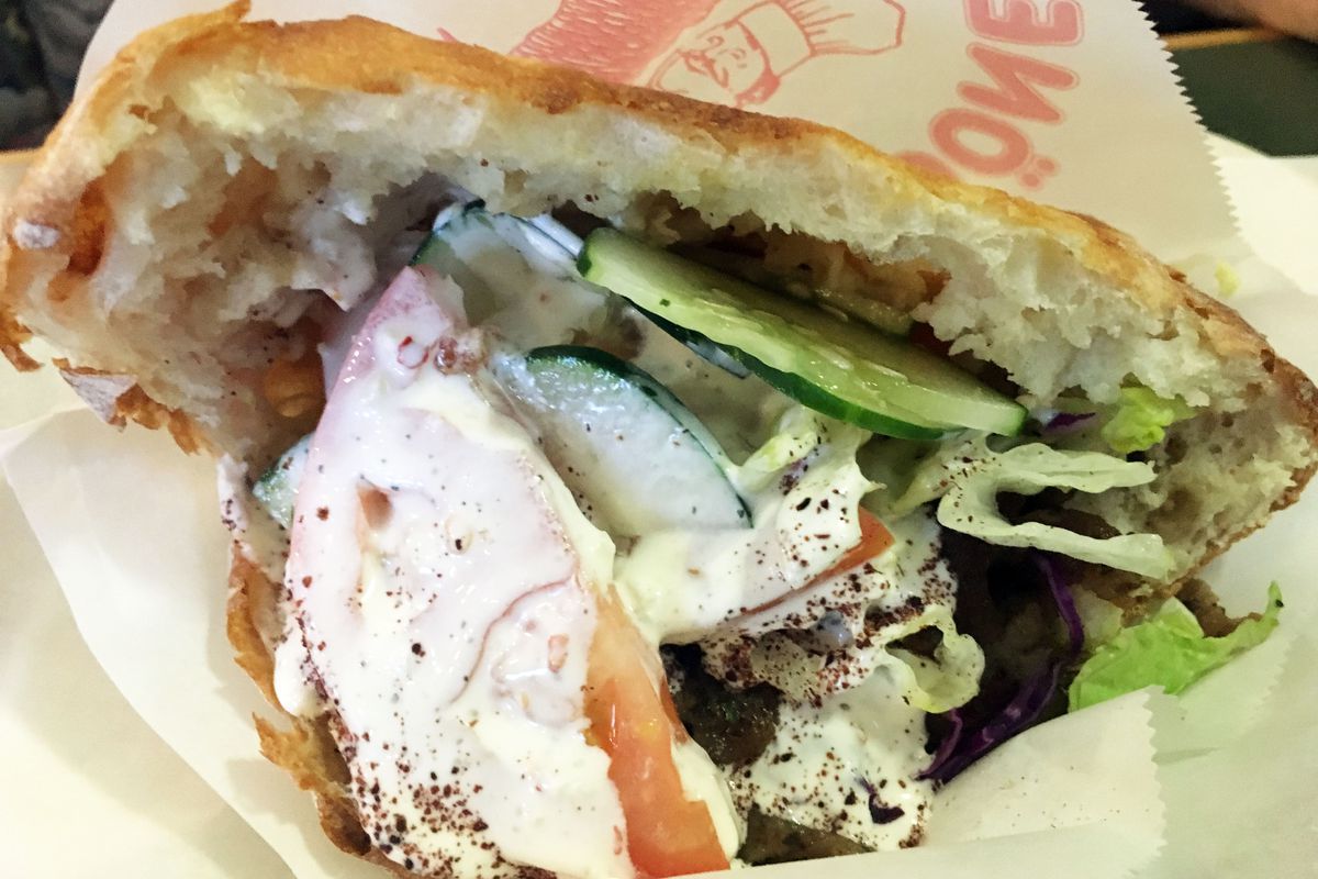 A close up of a döner sandwich with veggies and meat stuffed in a pocket of soft white bread and drenched with creamy white sauce.