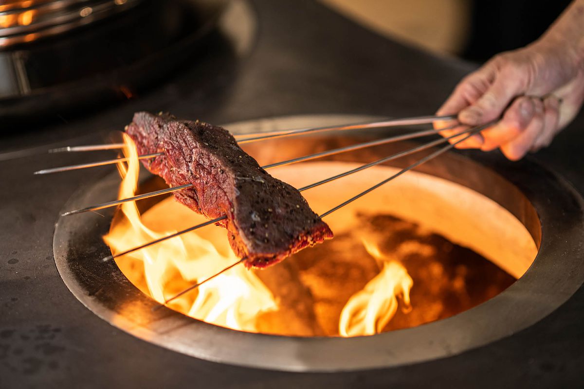 A hand holds metal skewers that run through a piece of red meat over an open flame at a restaurant.