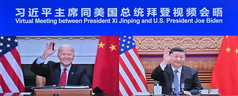 A screen capture from a video meeting is captioned in Chinese and English, reading “Virtual Meeting between President Xi Jinping and US President Joe Biden,” over side-by-side screens of Biden and Xi. Each is waving to the camera with a smile and each has one American and one Chinese flag flanking them as they sit at desks.