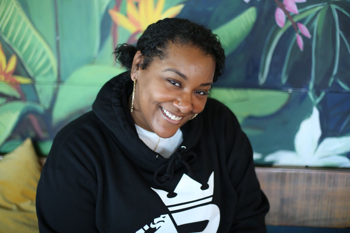 Sierra Carter, wearing a black sweatshirt with a bright white design on it. sitting in front of a green rainforest mural and smiling at the camera. 