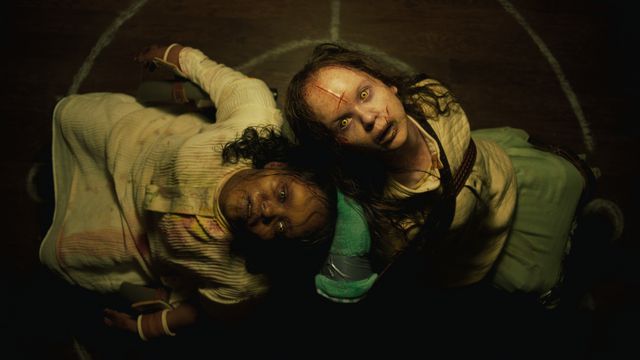 Two possessed, scarred and bruised children sit back to back on the floor and glare at the camera above them in The Exorcist: Believer