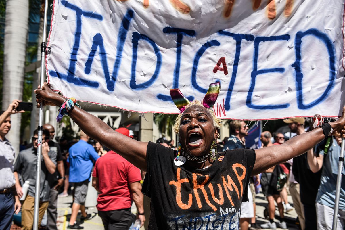 A Black woman wearing a shirt with anti-Trump writing on it holds a large sign saying “Indicted” above her head.