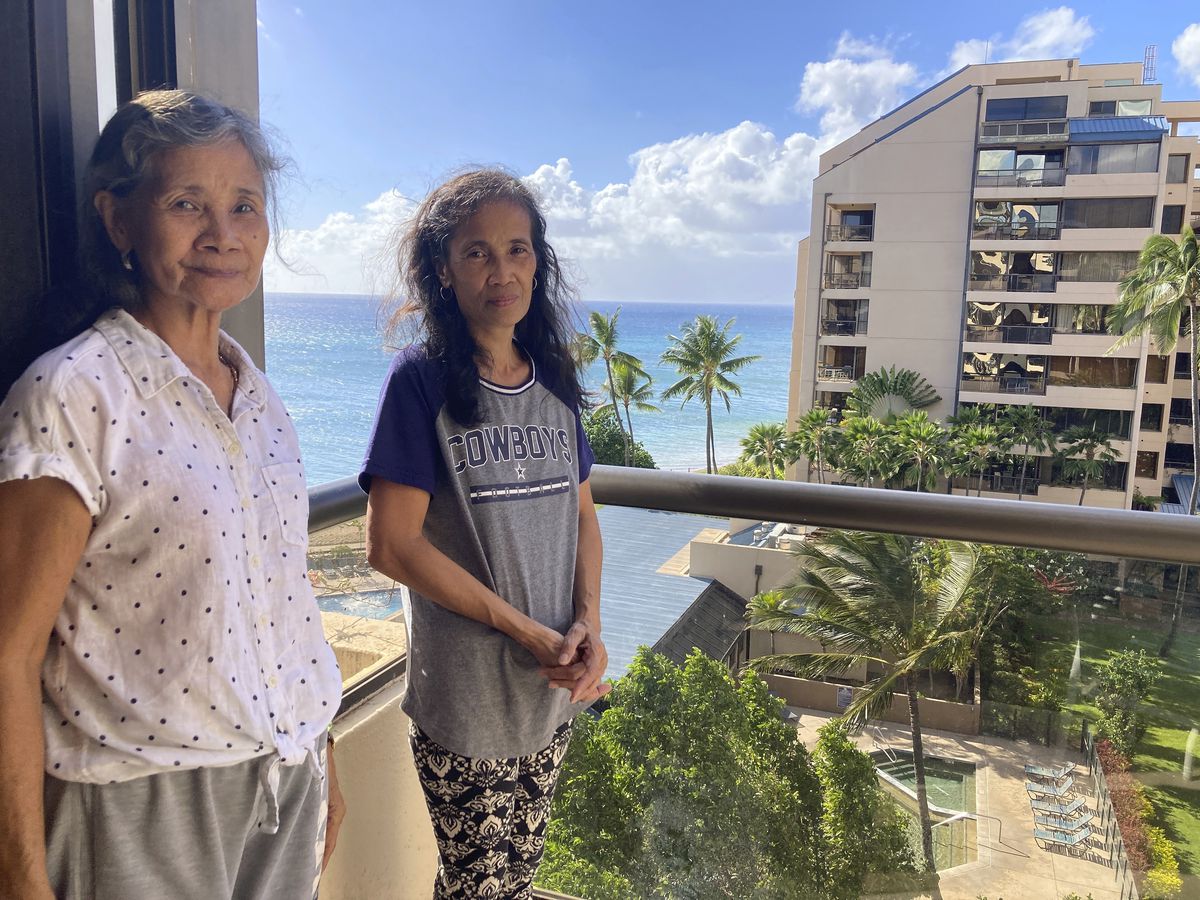 Two women stand on a hotel balcony with the ocean and palm trees in the background.
