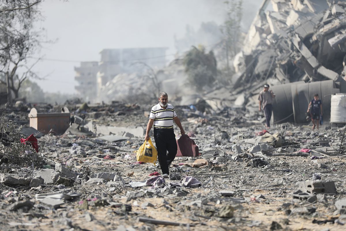 A man carrying two bags walks through piles of rubble and debris. The air is dusty and grayish. 