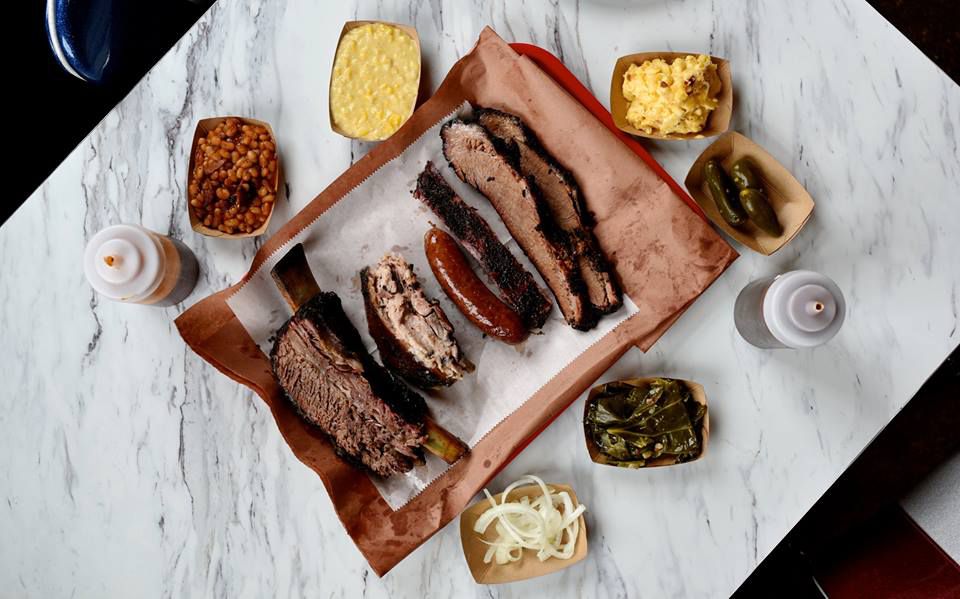 A meat tray with brisket, sausage, a beef rib, and pork ribs, surrounded by sides, including mac and cheese, creamed corn, beans, barbecue sauce, and trays of jalapenos and onions.
