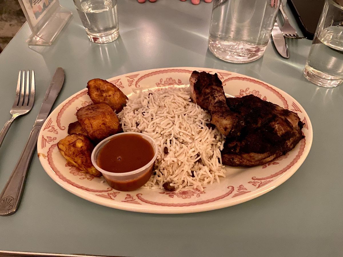 A white plate with red outlining is filled with plantains, white rice, jerk chicken and sauce. 