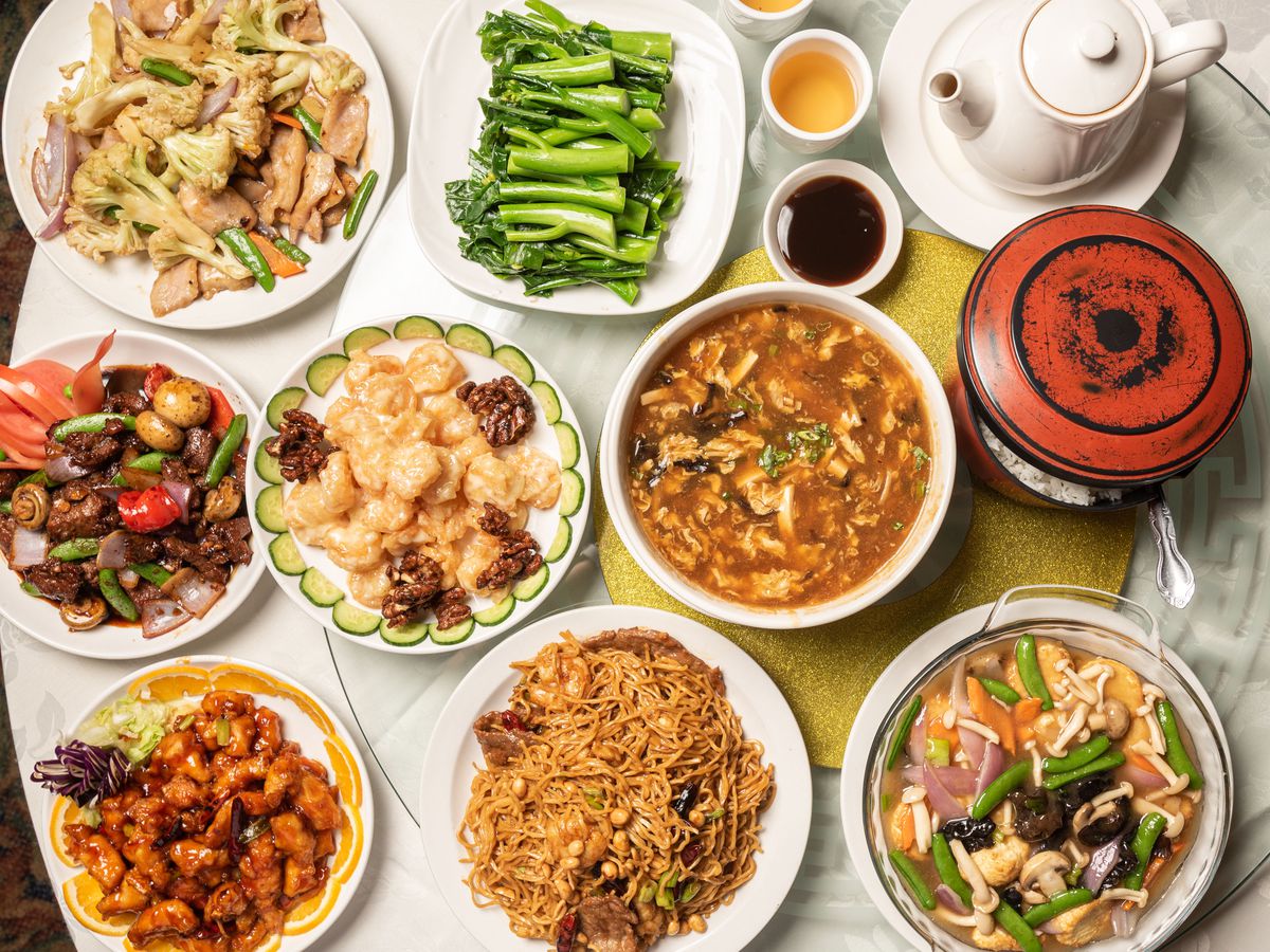 A table top brimming with Chinese food including vegetables, honey walnut shrimp, hot and sour soup, and more.
