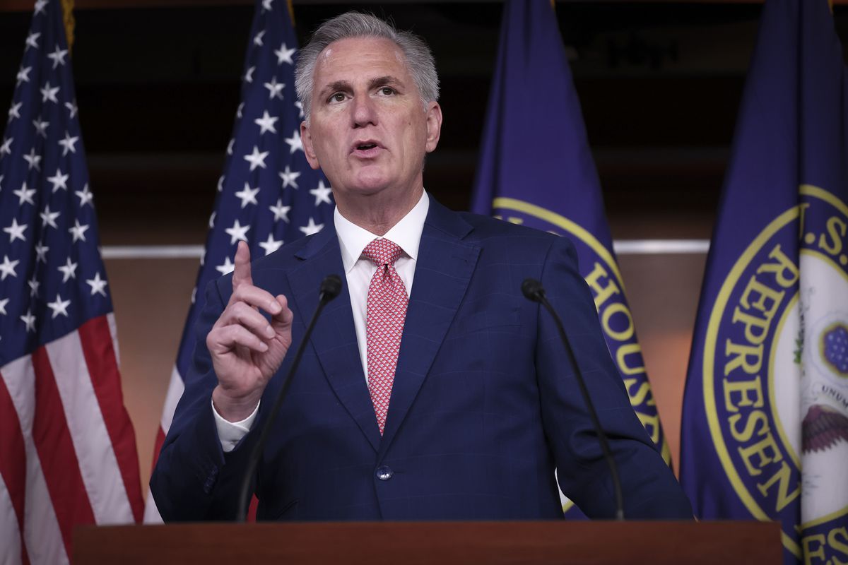 Kevin McCarthy, in a navy blue suit and red-patterned tie, points upward while speaking. A wooden podium is in front of him and behind him are several official flags.