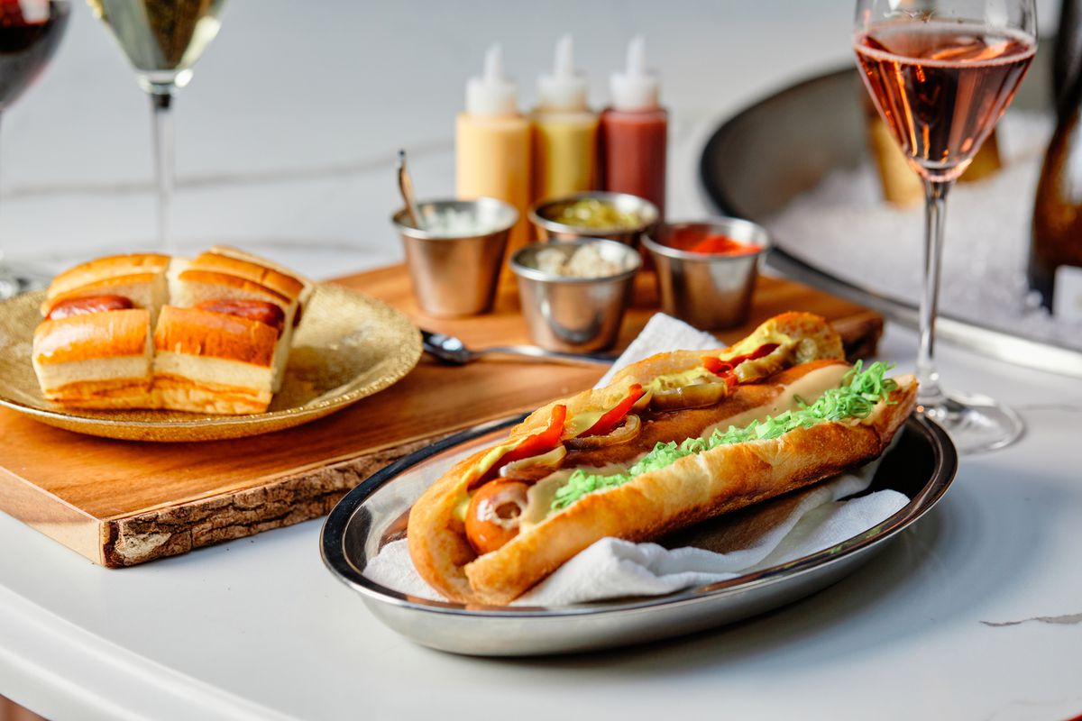A fancy-looking hot dog sits on a silver platter with glasses of wine in the background.