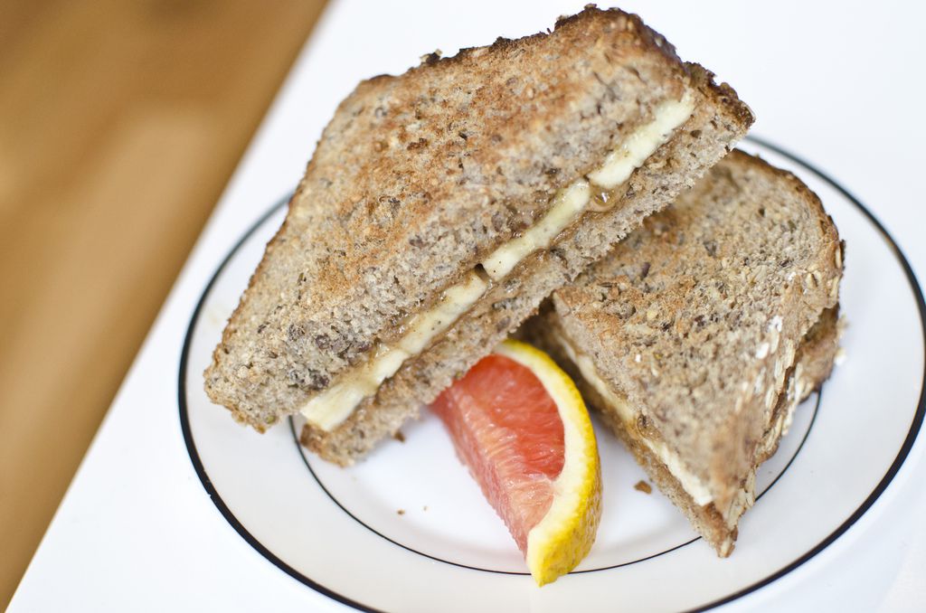 A sandwich with thick bread, banana, and peanut butter is slice diagonally and stacked on a white plate with a dark rim. There’s a small slice of grapefruit on the plate, and a wooden floor is visible in the background.