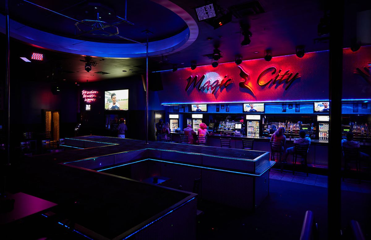 The neon-lit interior of Magic City features an H-shaped stage, a long bar with multiple tvs, and a window through which guests can order wings.