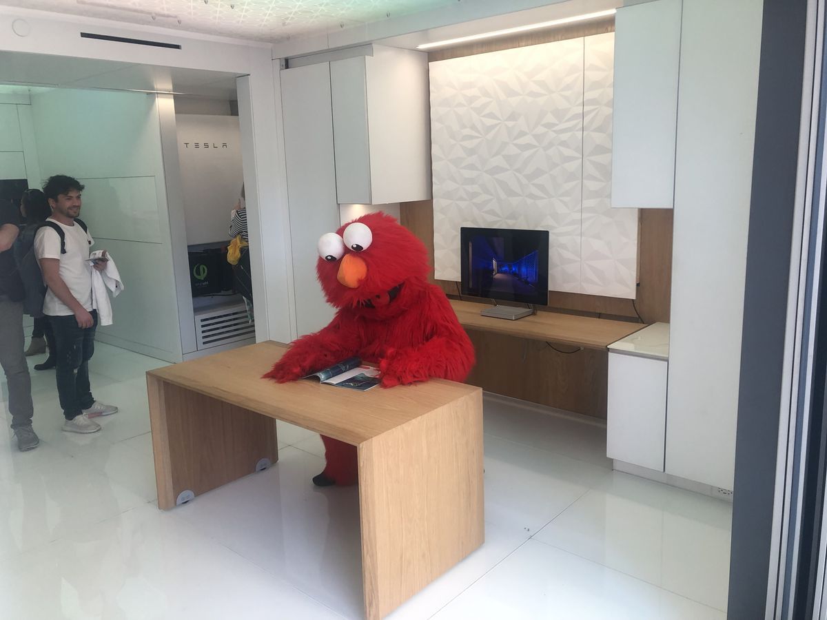 The interior of Futurehaus in Times Square. The Sesame Street character Elmo sits at a wooden table. The floors, walls, and ceiling are white. People look at Elmo and smile.